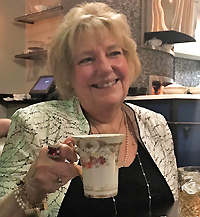 Genny Moir, an older blonde woman smiling and holding a teacup